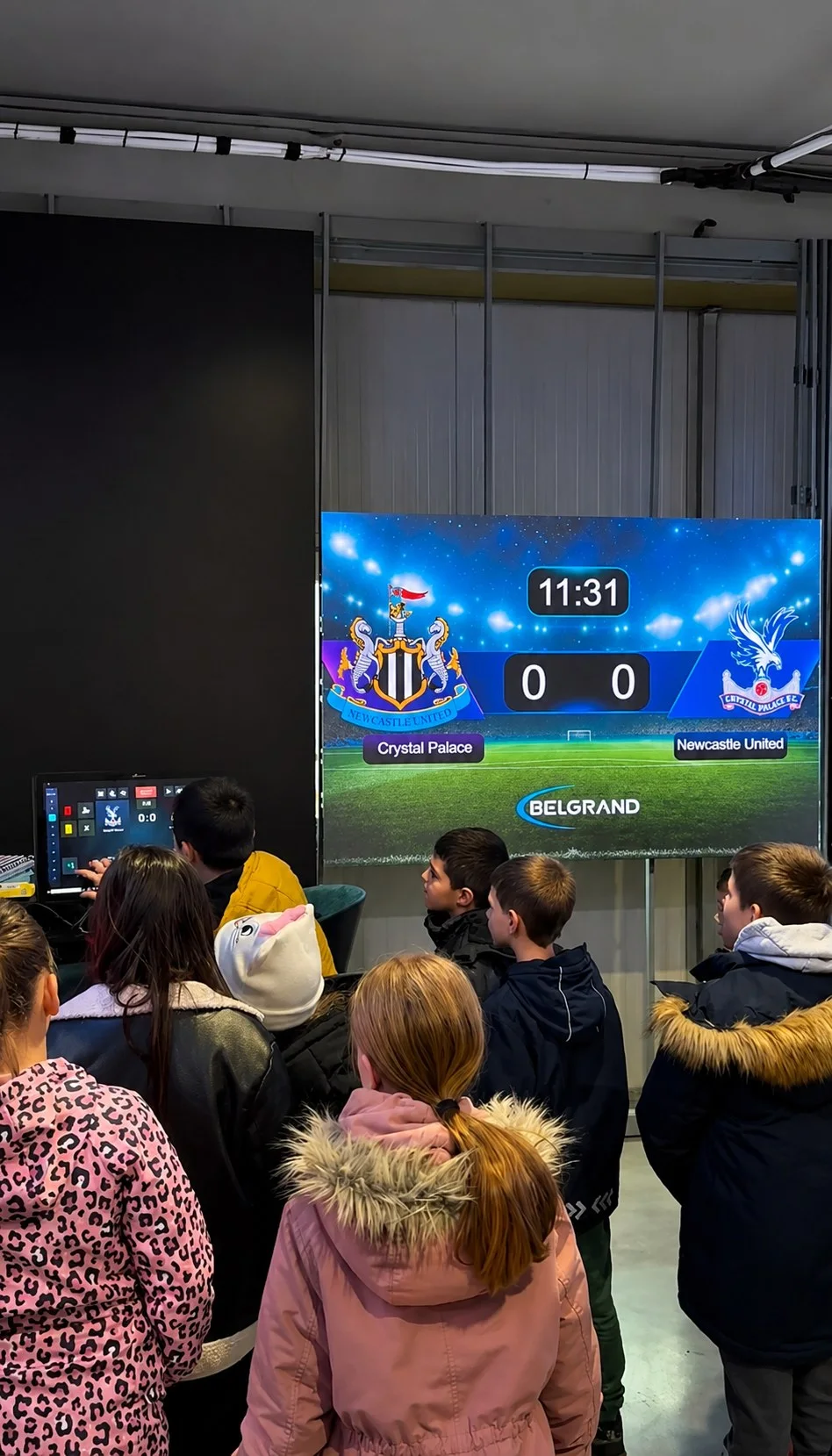 A group of children watching a live match score on a BELGRAND LED board, with the ScoreMaster control screen in the foreground